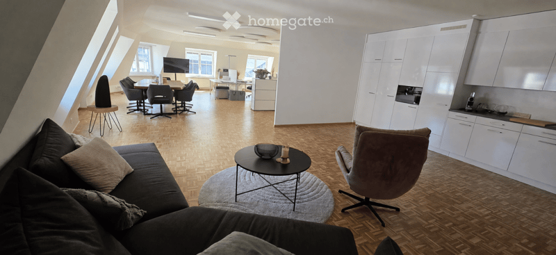 A spacious, modern living room with a dark grey sofa, coffee table, and a round side table. The room features a kitchen area with white cabinets and a dining area with a table and chairs. Large windows allow natural light to fill the space.