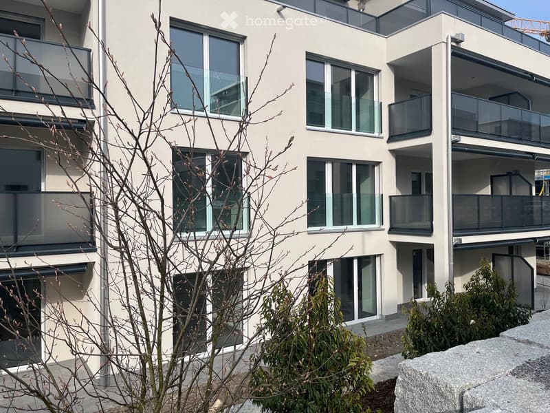 Modern multi-story residential building with large windows and balconies, surrounded by small trees and shrubs.