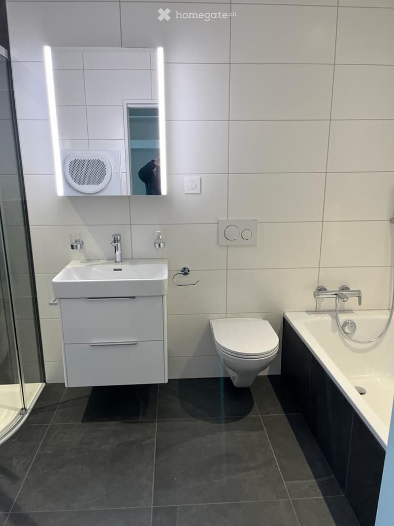 Modern bathroom with white tiles, dark floor, sink, toilet, bathtub, and illuminated mirror.