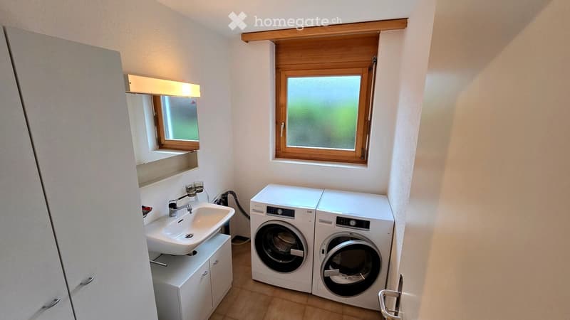 Bathroom with a sink, mirror, and a pair of stacked washing machine and dryer units. The room has two windows with wooden frames and a built-in light above the mirror.