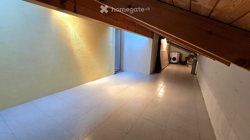 Empty hallway with tiled floor, exposed wooden beams on the ceiling, and a small room visible at the end containing a washing machine and other items.