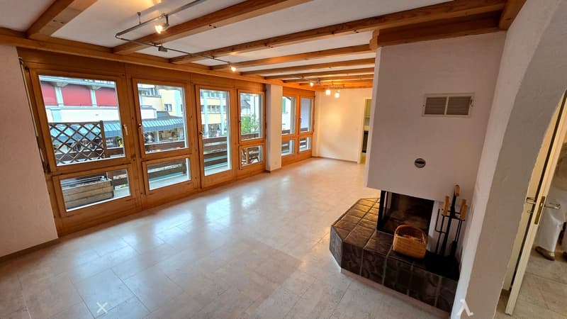 Empty room with wooden beams on the ceiling, large windows with wooden frames, tiled floor, and a fireplace with a small basket and fireplace tools.