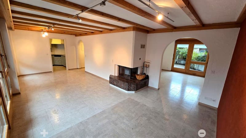 Bright, open living space with exposed wooden beams, tiled floor, fireplace, and large windows leading to an outdoor area.