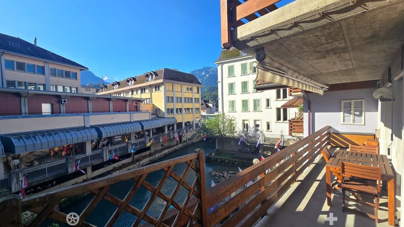 Balcony overlooking a canal with surrounding buildings and mountains in the background, featuring a wooden table and chairs.