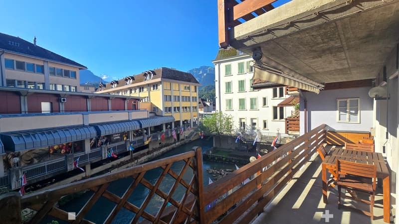 Balcony overlooking a canal with surrounding buildings and mountains in the background, featuring a wooden table and chairs.