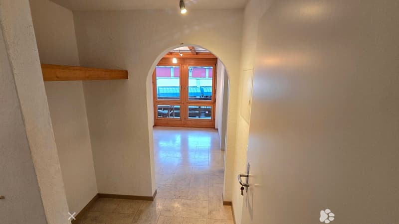 A well-lit hallway with beige tiled flooring and light-colored walls, featuring an arched doorway leading to another room with wooden doors and visible furniture.
