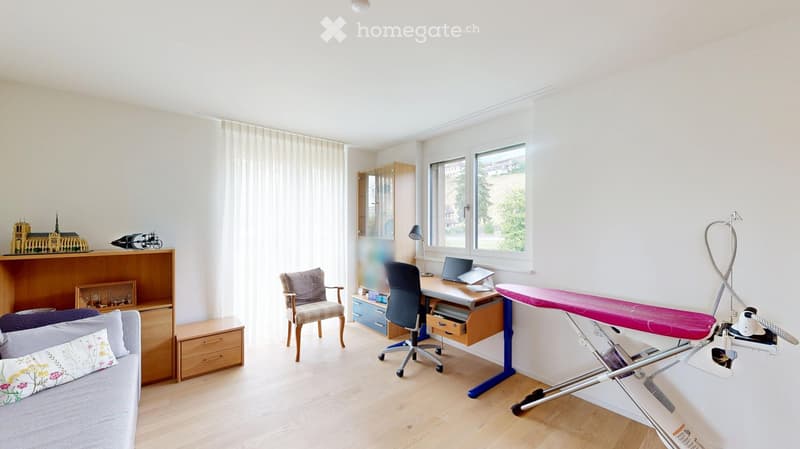 Bright room with wooden flooring, large windows, a sofa, a desk with a laptop, a chair, and an ironing board with an iron.