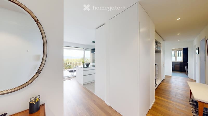 Bright hallway with wooden flooring, large round mirror on the wall, leading to a kitchen area and a balcony with a view.