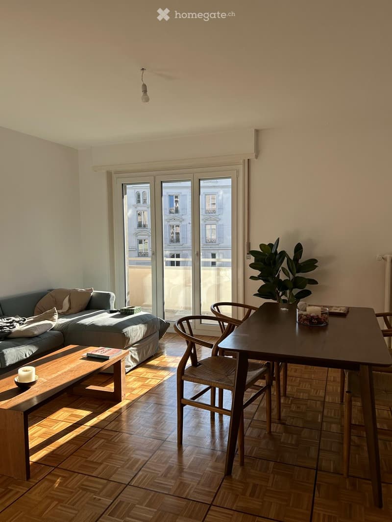 A modern living room with a grey sofa, wooden coffee table, dining table with chairs, and a potted plant near the window. The room features wooden flooring and large glass doors leading to a balcony.