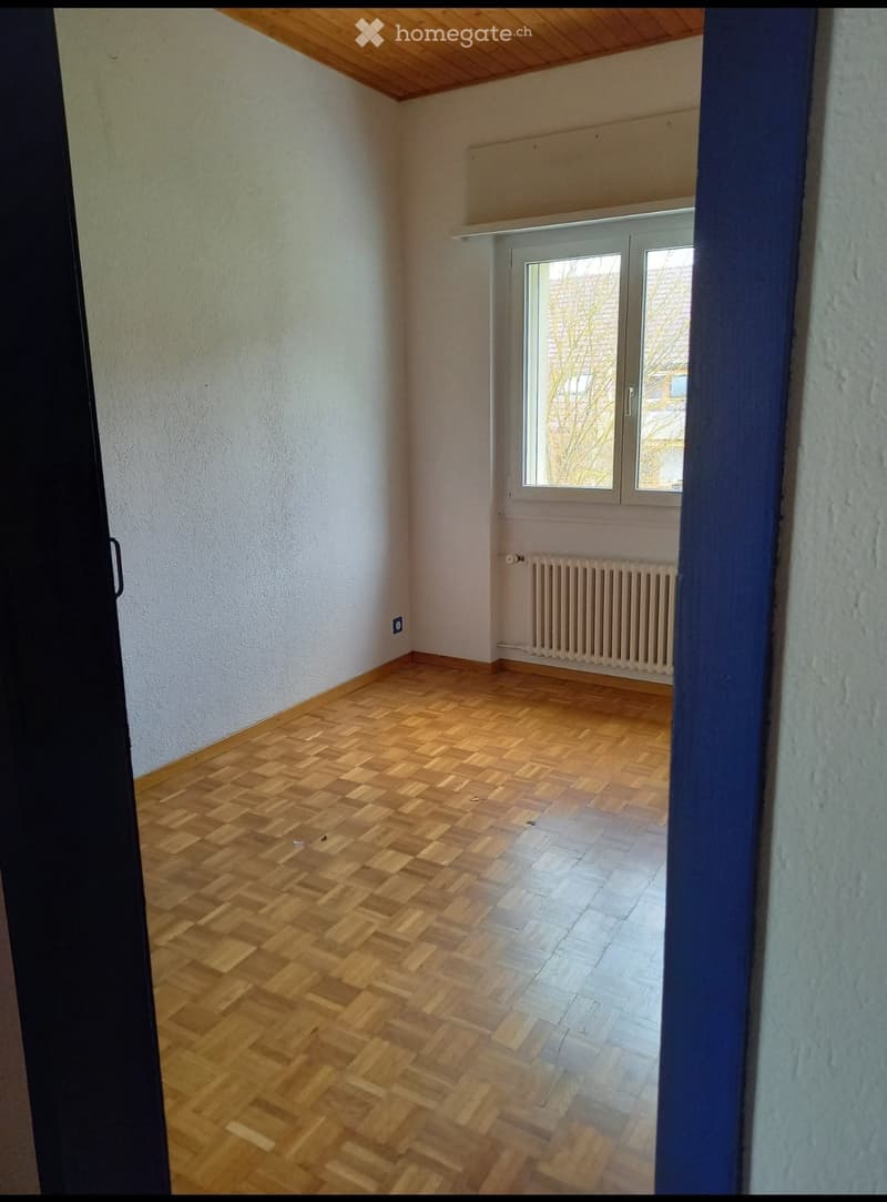 Empty room with wooden herringbone flooring, white walls, and a window with a radiator below it.
