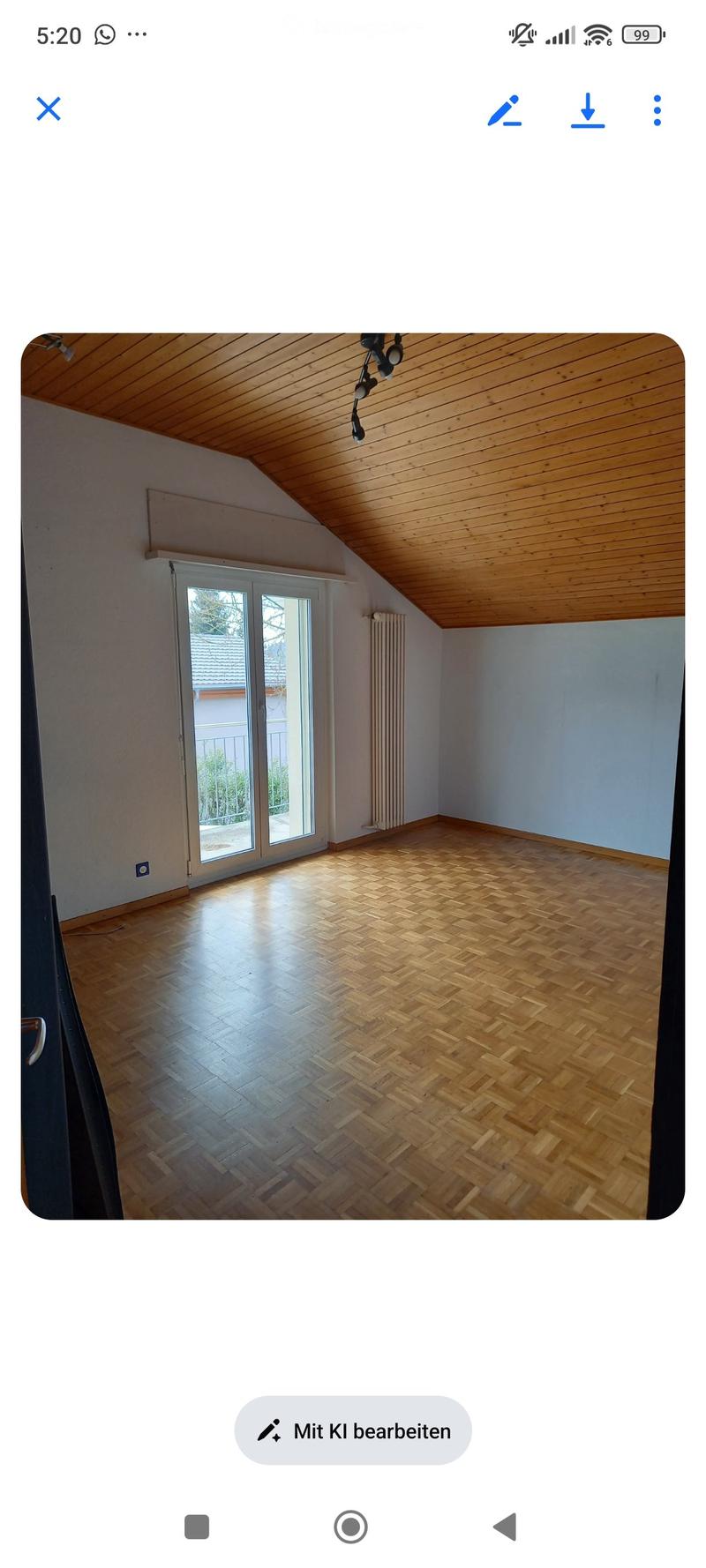 Empty room with wooden herringbone flooring, white walls, and a large window with a view of the outside. The ceiling is slanted with wooden paneling.