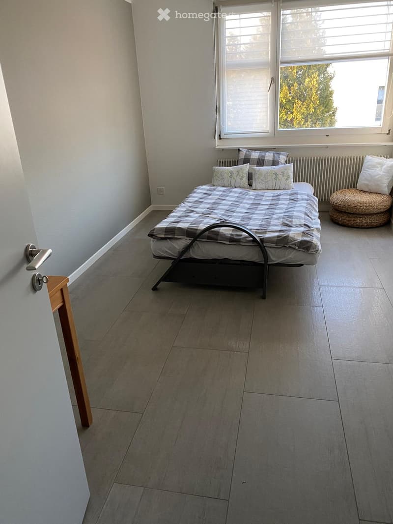 A modern bedroom with a grey tiled floor, a metal-framed bed with a checkered duvet, pillows, and a small wicker stool near the window.