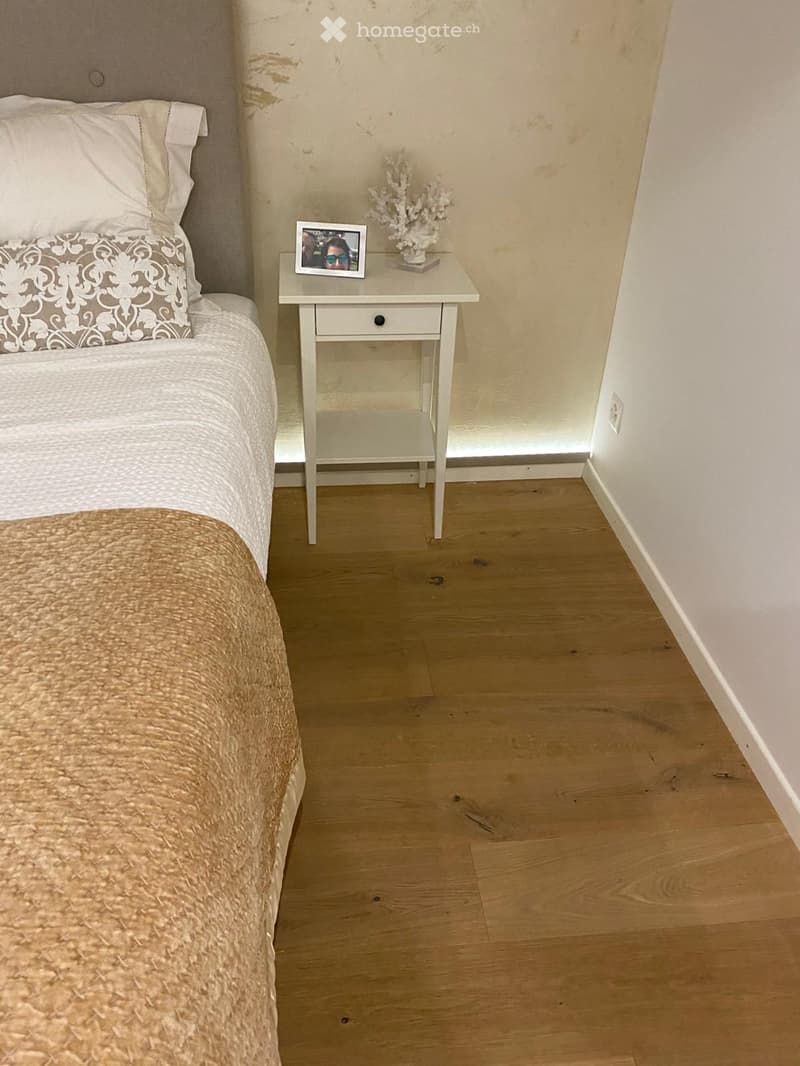 A cozy bedroom with a neatly made bed, a nightstand with a picture frame and decorative item, and light wooden flooring.