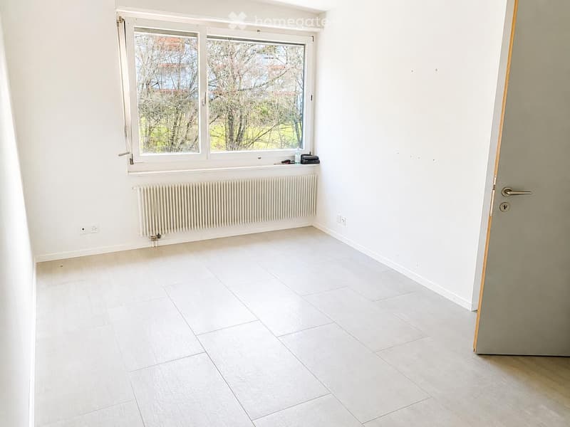 Empty room with white tiled flooring, white walls, a large window with a view of trees, a radiator, and a door.