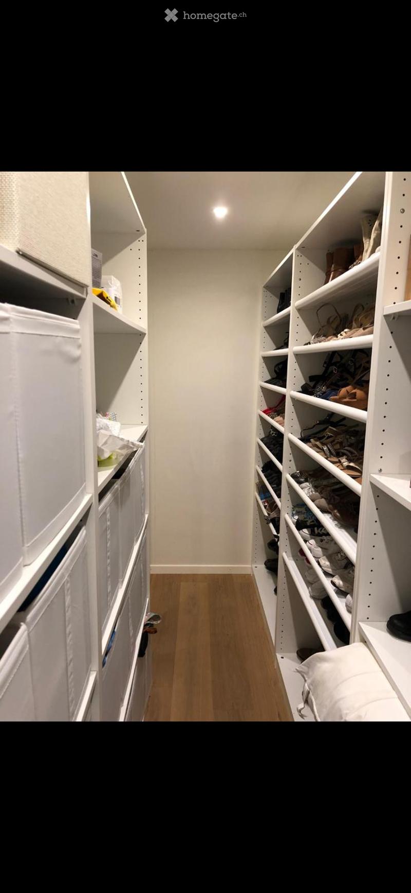 Spacious, well-lit closet with white built-in shelving units on both sides, containing neatly organized clothing, shoes, and storage boxes. The floor is light wooden, and the walls are white.