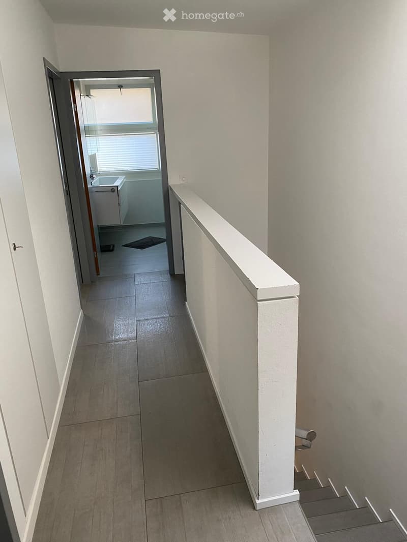 A narrow hallway with tiled flooring, white walls, and a staircase leading upwards. An open doorway leads to a small bathroom with a sink, toilet, and bathtub.