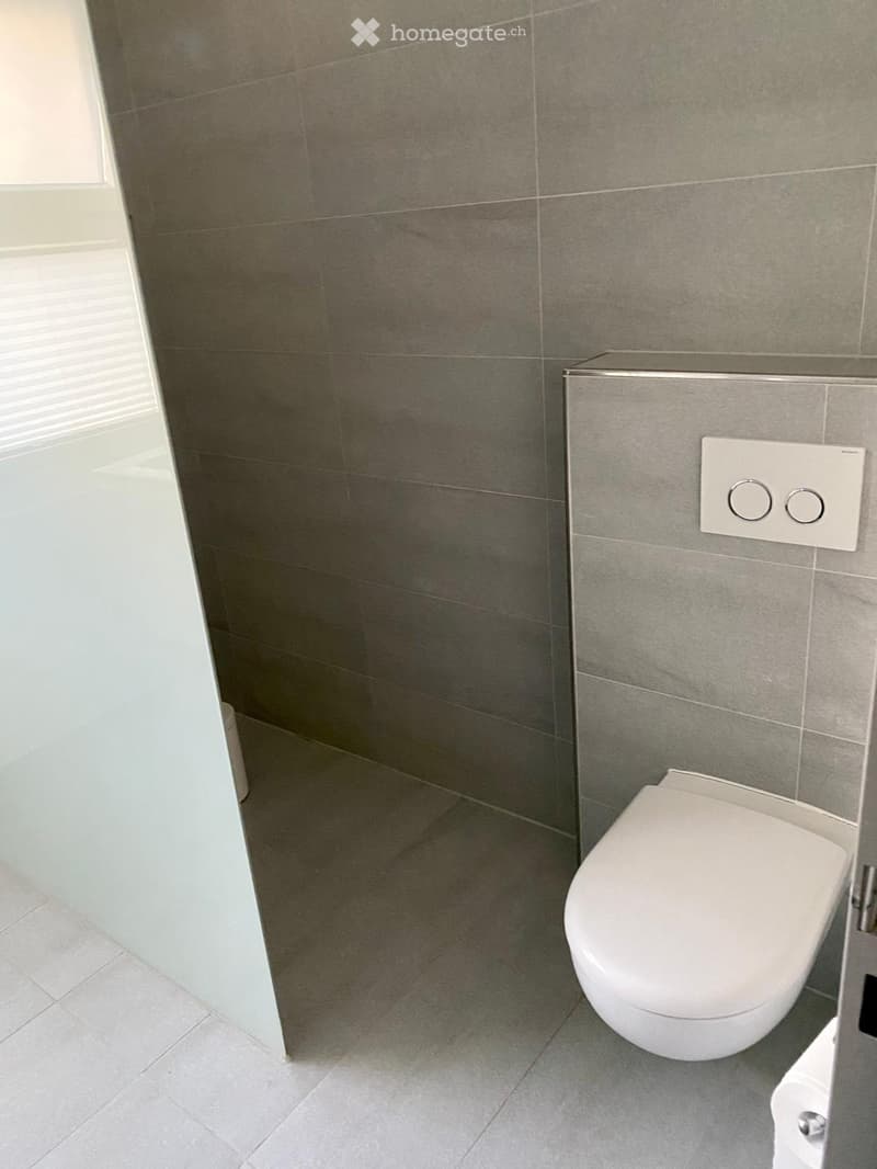 Modern bathroom with grey tiled walls and floor, white toilet, and a control panel on the wall.