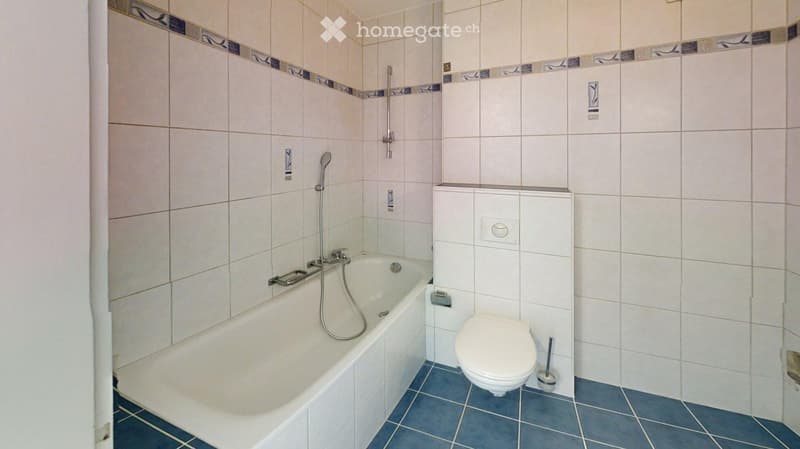 Bathroom with a bathtub, toilet, showerhead, and faucet. The walls are tiled with a decorative border, and the floor is blue tiled.