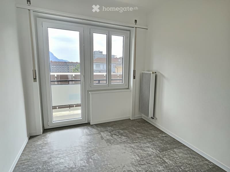 Empty room with grey tiled floor, white walls, large sliding glass door leading to a balcony, and radiators on both sides of the door.