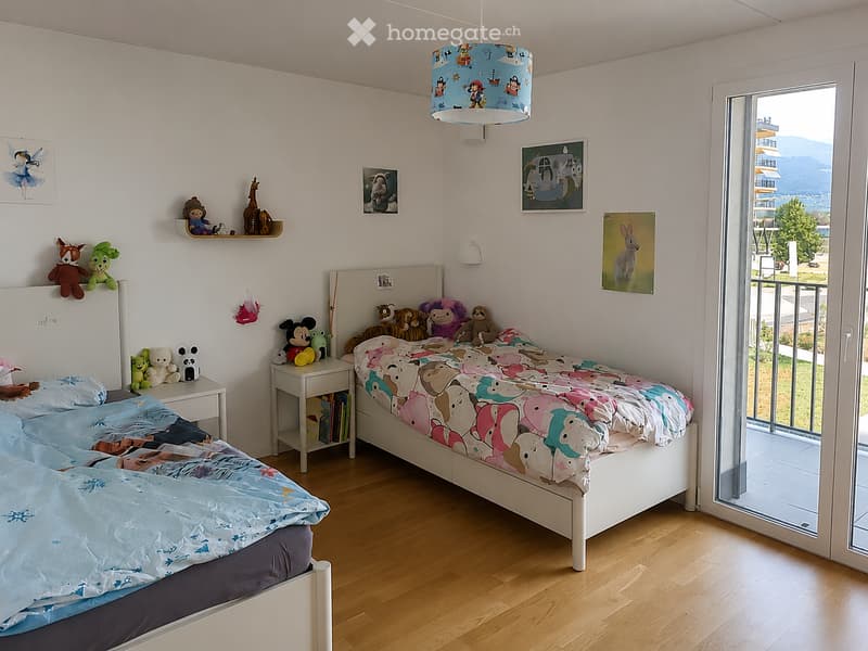 Bright, child-friendly bedroom with two single beds, colorful bedding, nightstand, toys, and artwork on the walls. Large window offers a scenic view.