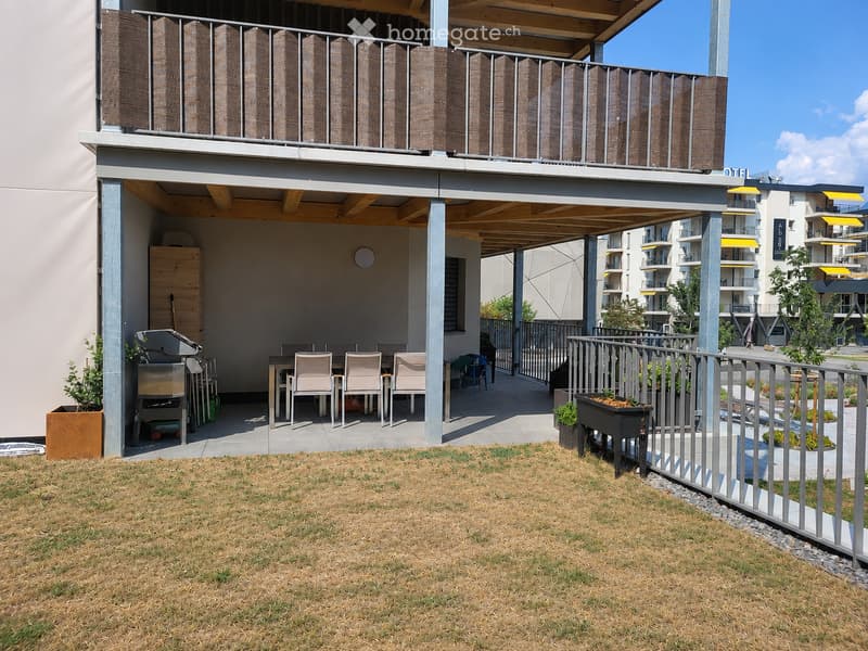 Balcony with a covered seating area, a grill, a table with chairs, and potted plants. The balcony is part of a modern residential complex with contemporary design elements.