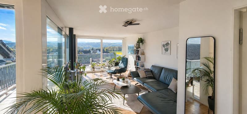Bright living room with large windows and a view, featuring a dark blue sofa, coffee table, armchair, and decorative plants.
