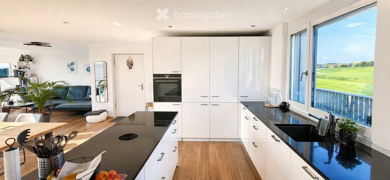 Modern kitchen and living area with white cabinets, black countertops, a large window with a scenic view, a sofa, a dining table, and various kitchen appliances.