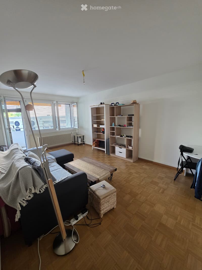 Living room with wooden flooring, large window, sofa with throw blanket, coffee table, ottoman, bookshelf, and floor lamp.