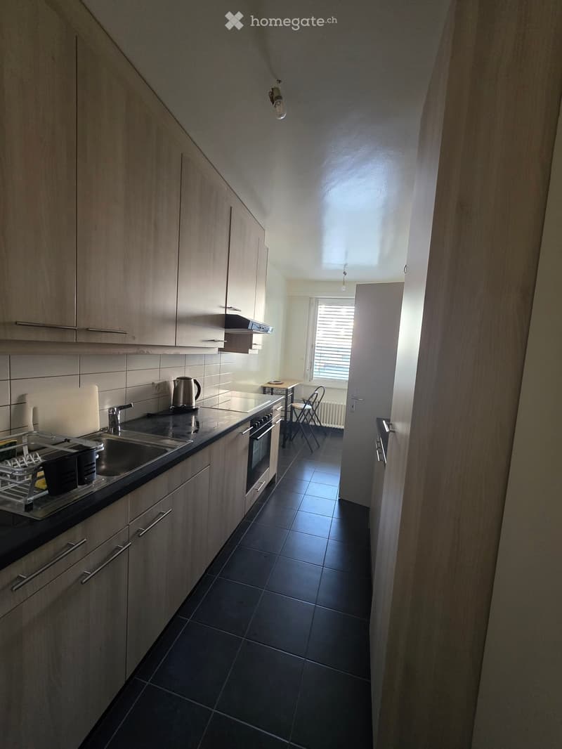 Modern kitchen with light wood cabinets, black tiled floor, stainless steel sink, oven, and small table with chairs in the adjacent area.