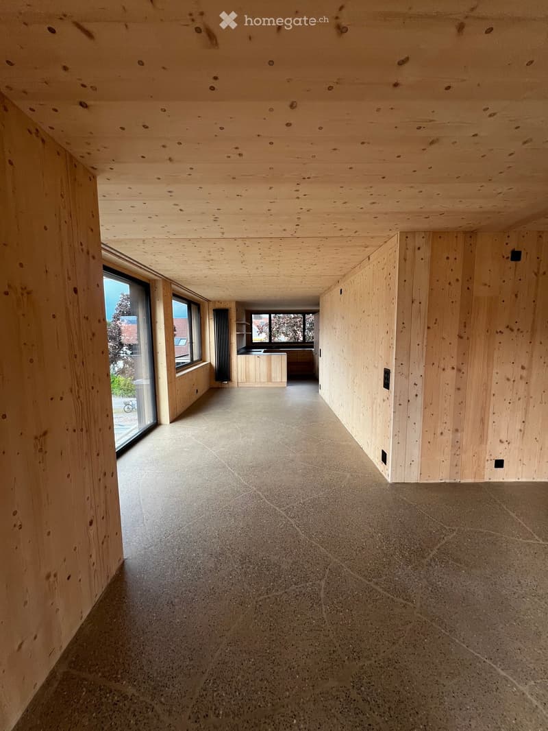 Empty wooden-paneled hallway with concrete floor, large windows, and visible kitchen area in the background.