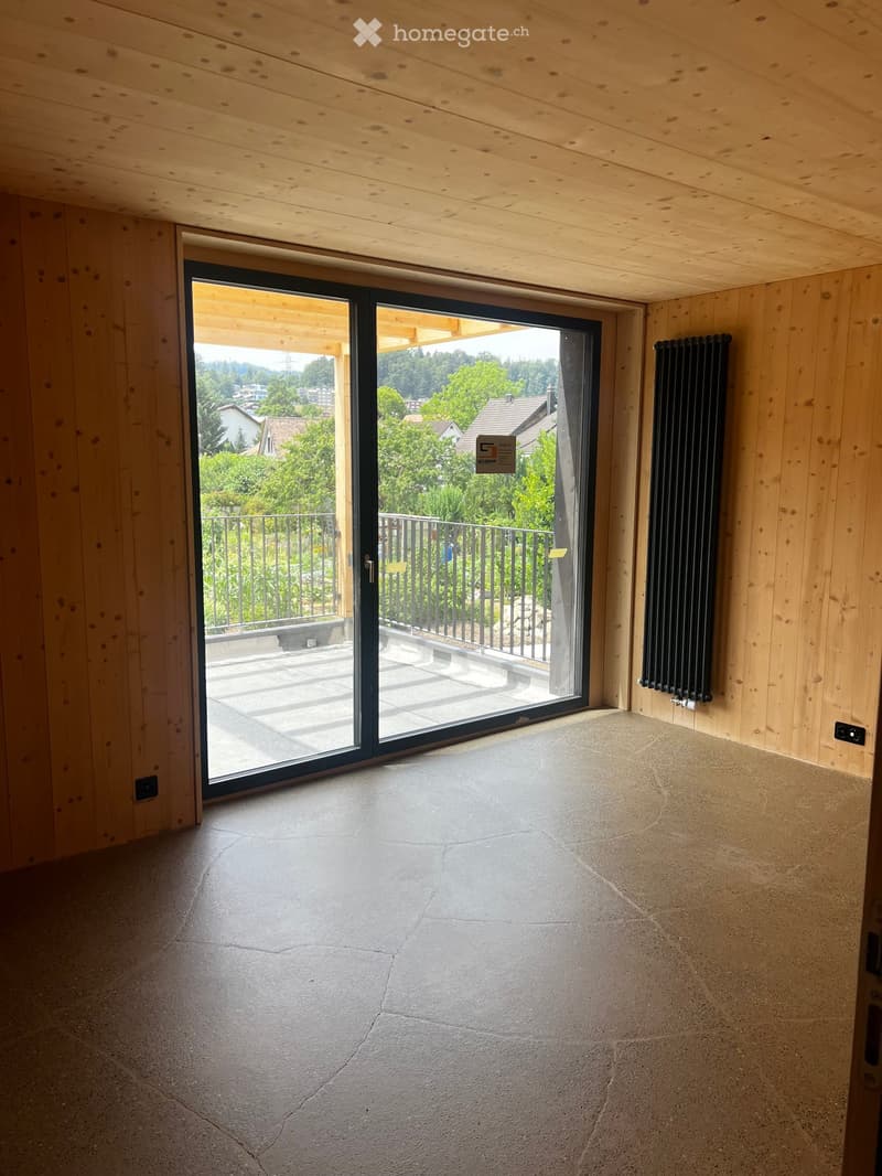 Empty room with wooden walls and ceiling, large sliding glass door leading to a balcony with a view of greenery and houses.