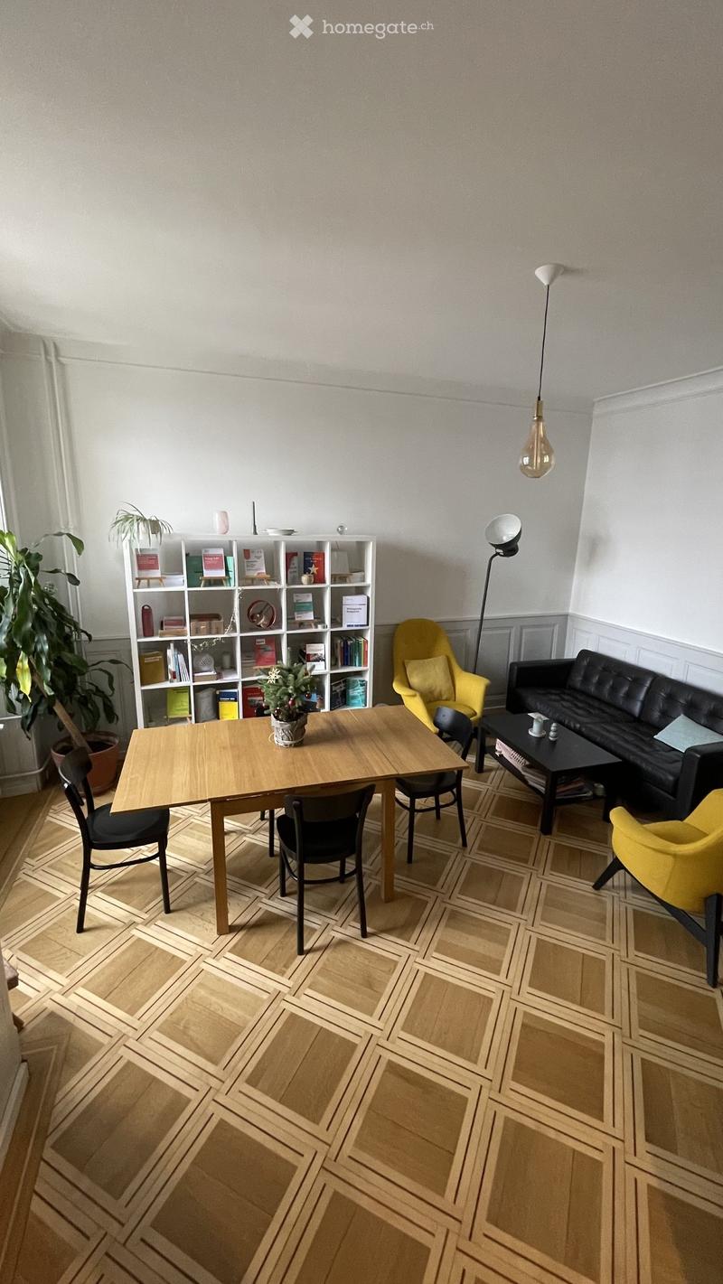 A modern living room with a wooden dining table, black chairs, a yellow armchair, a black leather sofa, a bookshelf, and a coffee table.