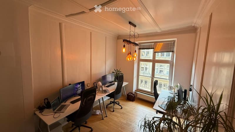 Bright, well-lit room with wooden flooring, two desks, multiple monitors, laptops, chairs, and decorative plants.