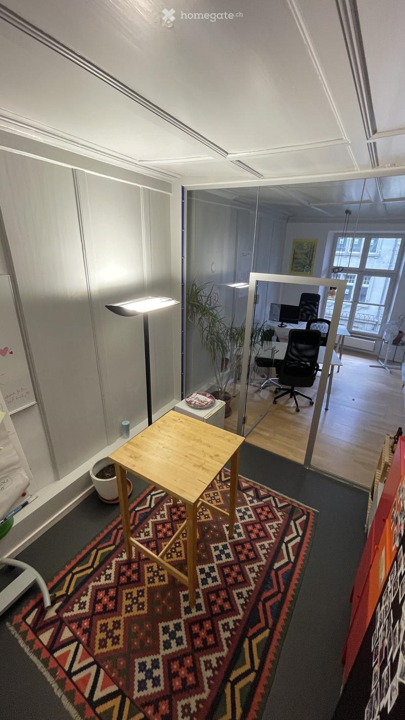 A modern office space with a small wooden table, chairs, a potted plant, and a large glass partition leading to another room with desks and office chairs.