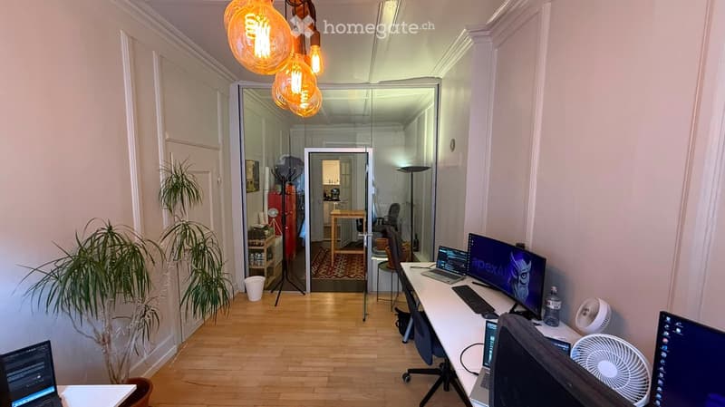 A modern, well-lit workspace with a large desk, multiple monitors, a laptop, and a comfortable chair. The room features wooden flooring, white walls, and decorative lighting. A potted plant adds a touch of greenery, and a small fan is placed on the desk f