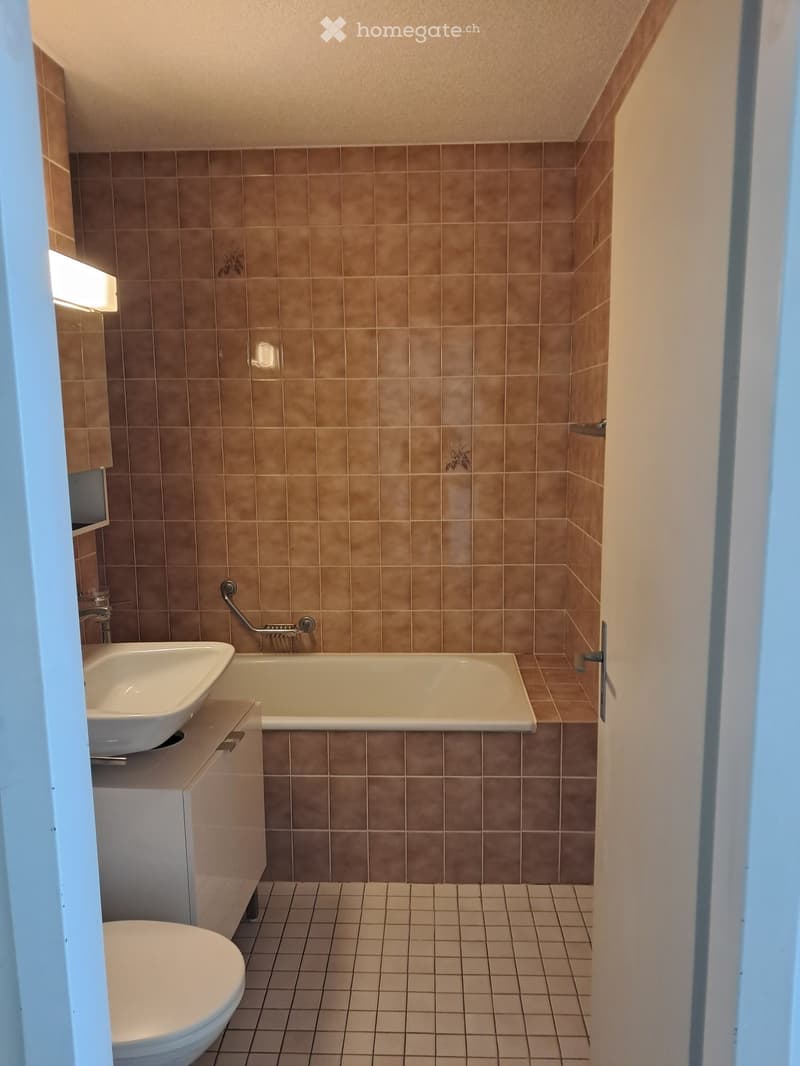 Bathroom with beige tiles, a bathtub, a sink, a toilet, and a small cabinet.