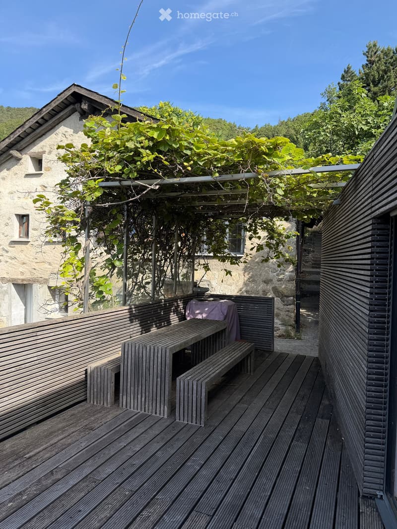 Wooden deck with benches and a small table covered with a pink cloth, surrounded by greenery and a stone building.