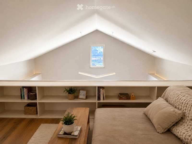Bright, airy bedroom with slanted ceilings and large window offering a scenic view. Features a bed with a textured blanket and pillows, a wooden side table with a plant and magazines, built-in shelving with books, baskets, and decorative items.