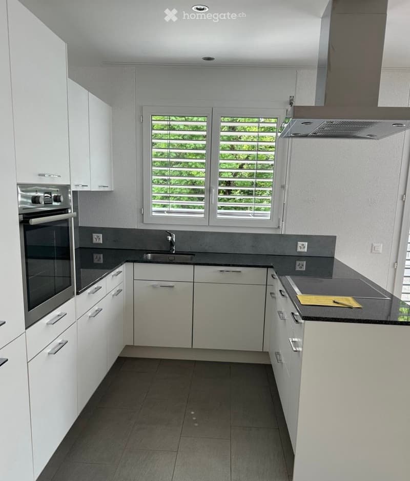 Modern kitchen with white cabinets, stainless steel oven, sink, and countertop. Features a large window with blinds and a dark tiled floor.
