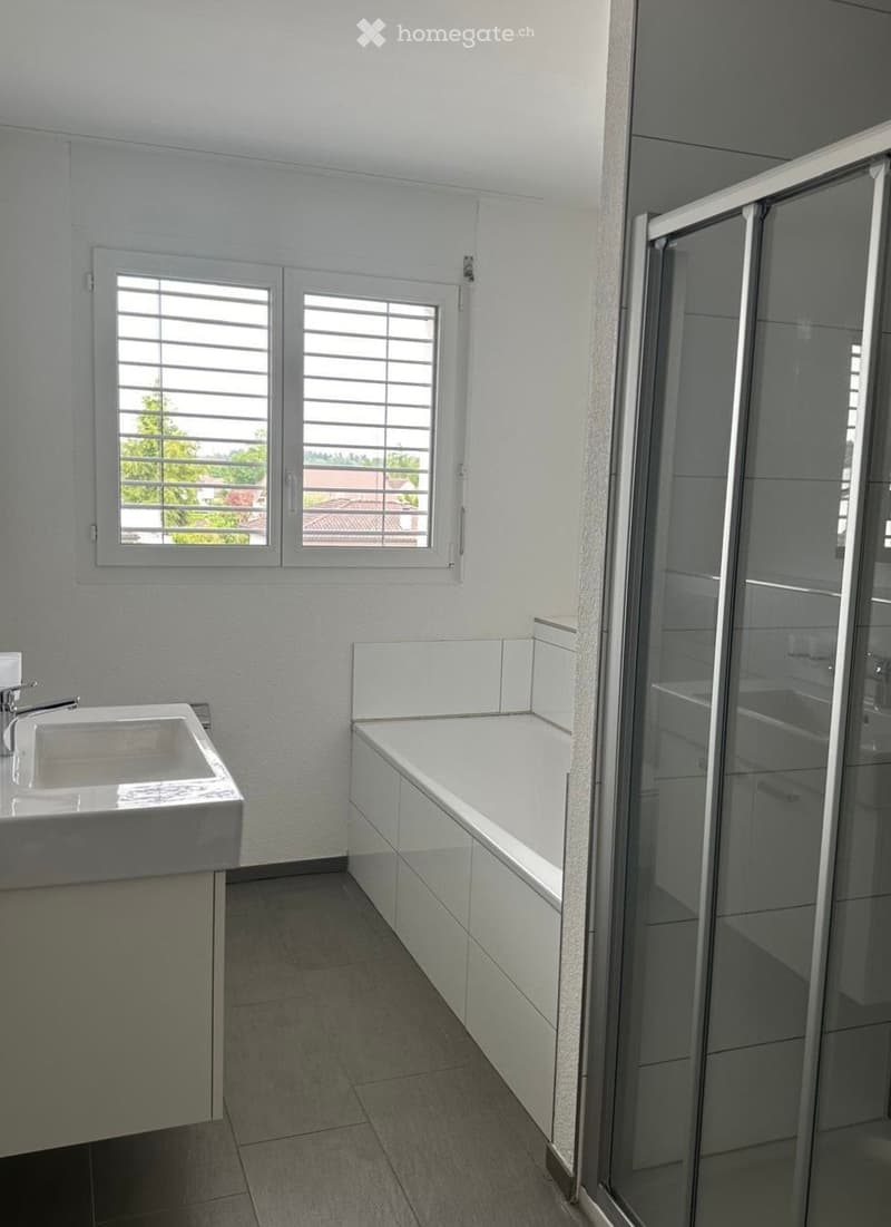 Modern bathroom with a white sink, bathtub, and glass-enclosed shower. The room features large windows with blinds and light-colored tiles.