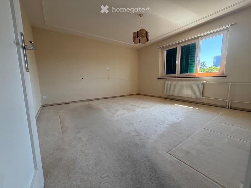 Empty room with beige walls, carpeted floor, large window with view, radiator, and chandelier.