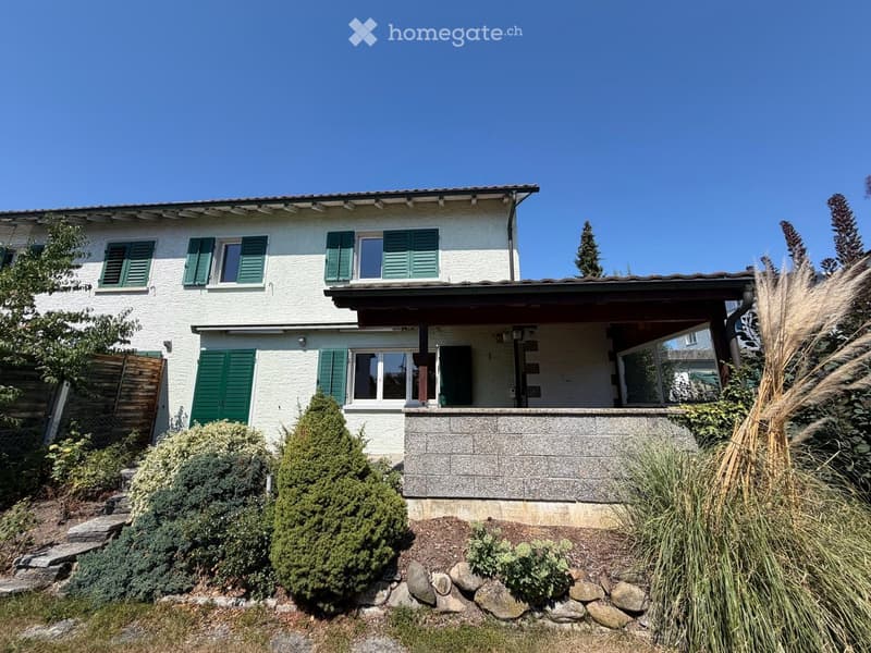 Two-story house with white exterior, green shutters, and a covered entrance area. The house is surrounded by well-maintained landscaping with shrubs, trees, and a stone pathway leading to the entrance.