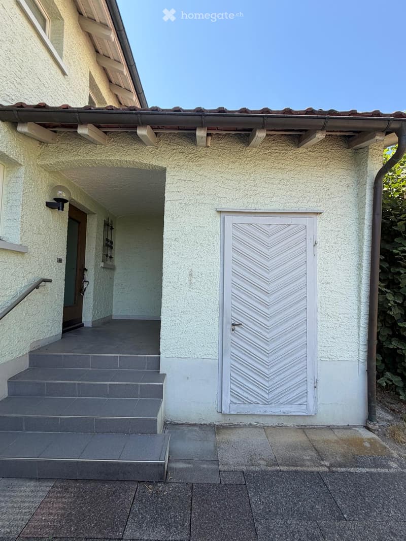 Exterior view of a two-story house with a raised ground floor, featuring a covered entrance with steps, a white door with a geometric pattern, and a small balcony area.