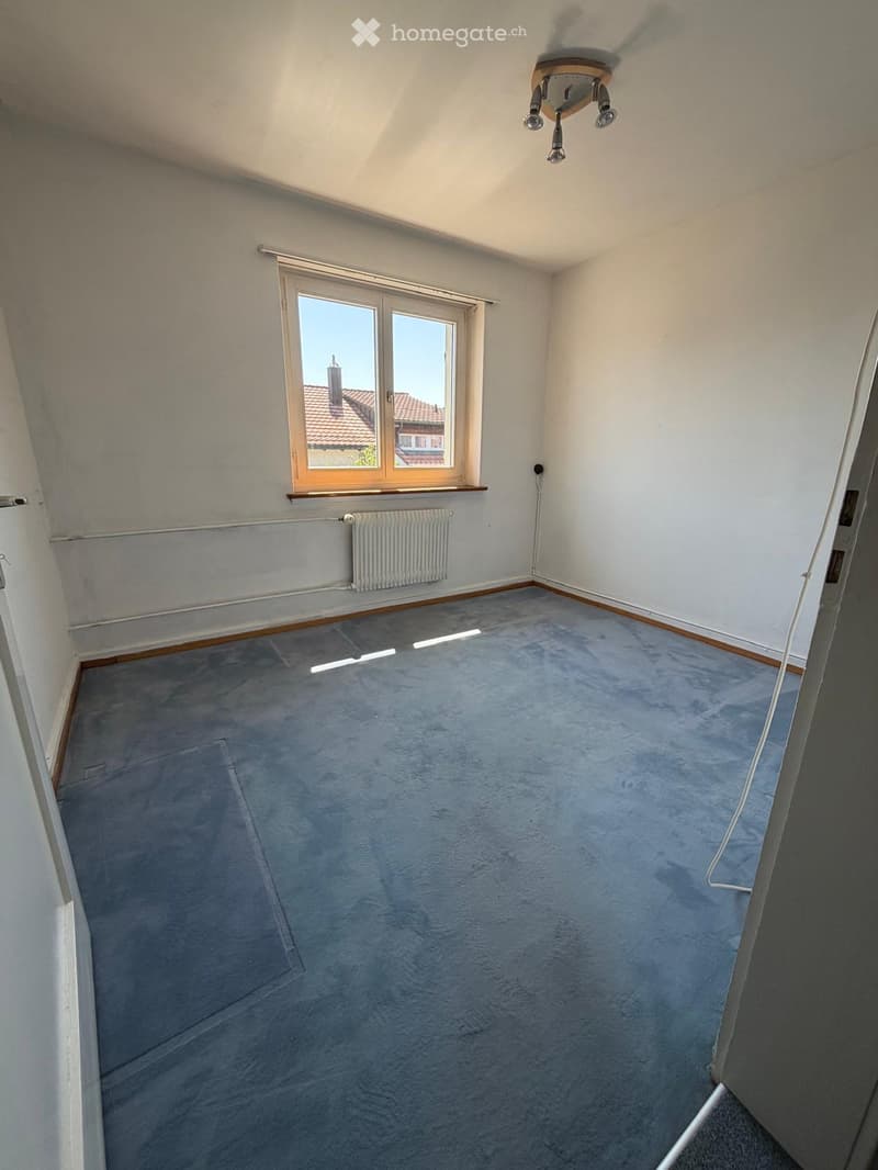 Empty room with grey carpet, white walls, large window with wooden frame, radiator, and ceiling light fixture.