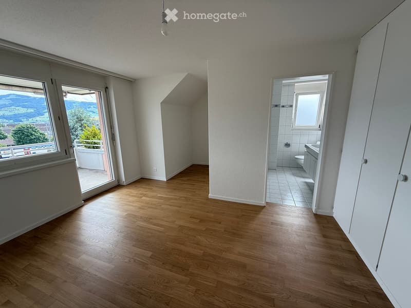 Empty room with wooden flooring, white walls, and large windows offering a scenic view. A bathroom is visible through an open doorway.