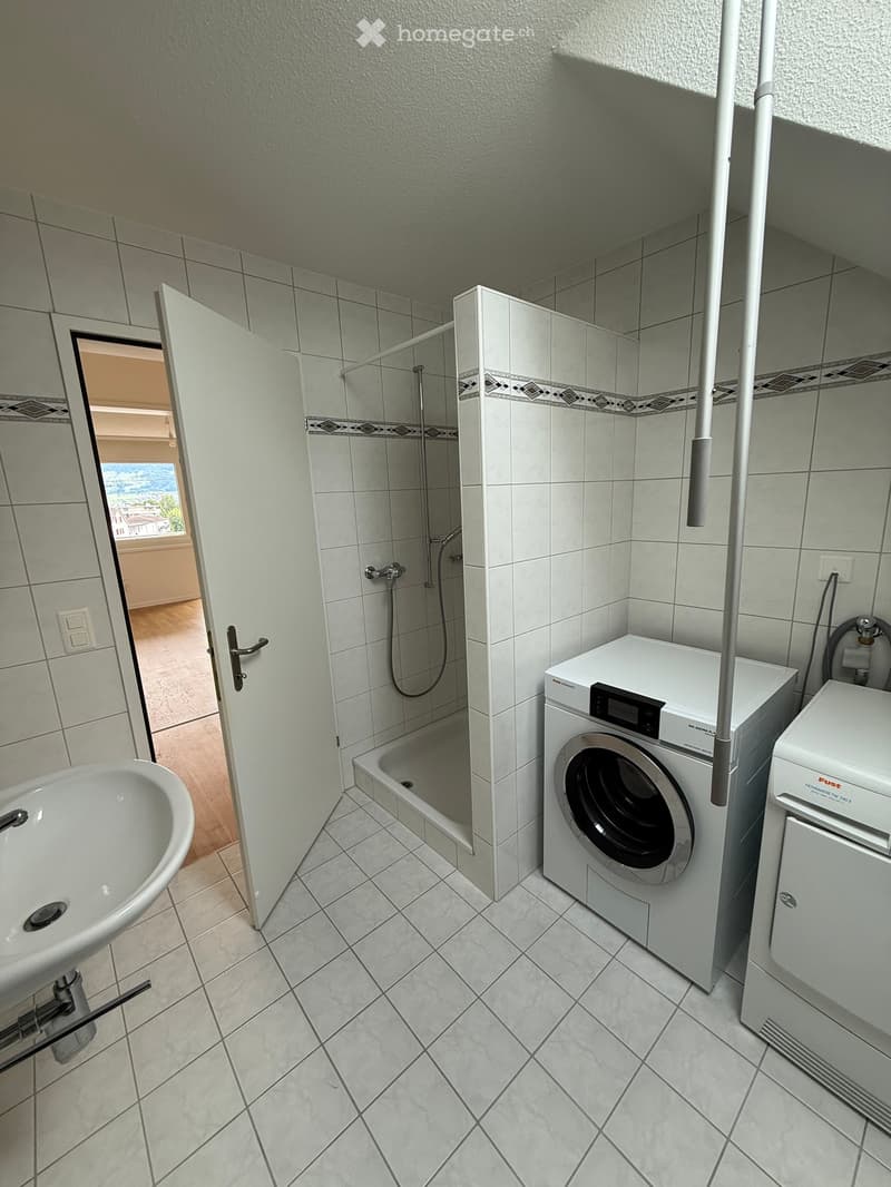Bathroom with tiled walls and floor, featuring a bathtub, shower stall, washing machine, and dryer. The shower has a glass door and a handheld showerhead.