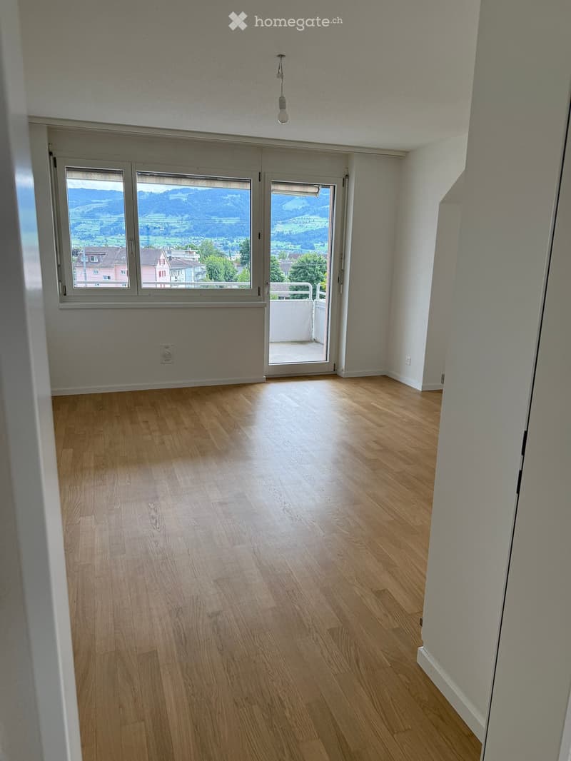 Empty room with wooden flooring, large windows, and a sliding glass door leading to a balcony.