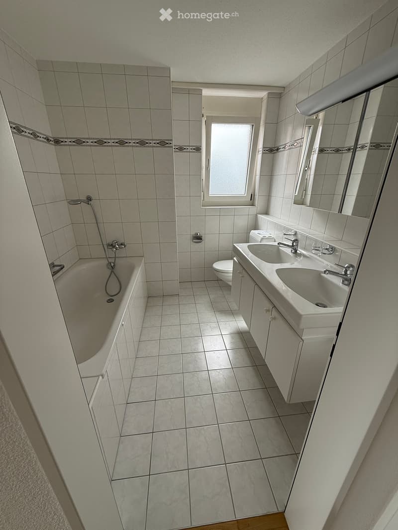 Modern bathroom with tiled walls and floor, featuring a bathtub, double sink, toilet, and a large mirror above the sink.
