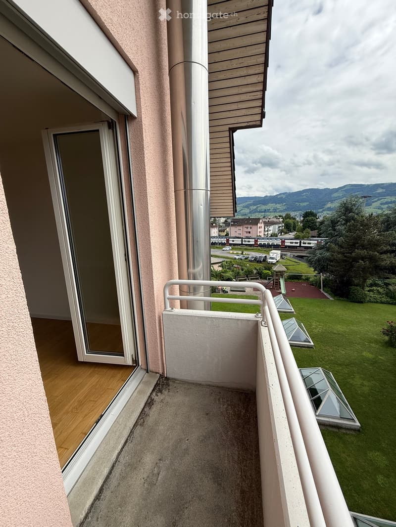 Balcony with wooden floor, white railing, and a view of a green lawn, playground, and distant mountains.