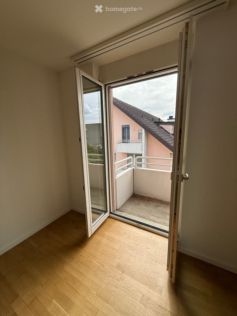 Open sliding glass doors lead to a small balcony with a concrete floor and white railing, overlooking neighboring buildings.